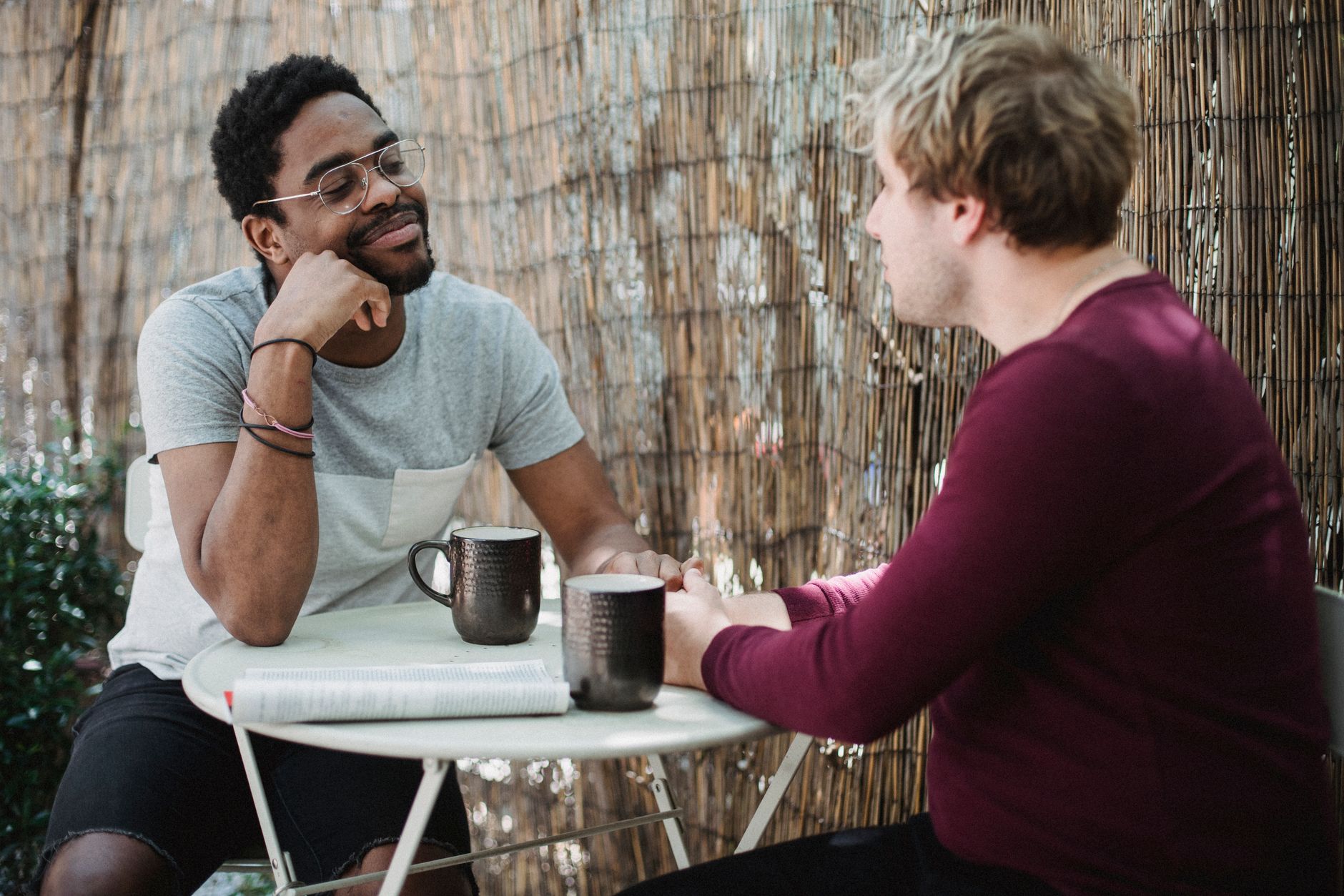 Deux hommes discutent en terrasse autour d’une petite table ronde, avec deux tasses et un journal devant eux.
