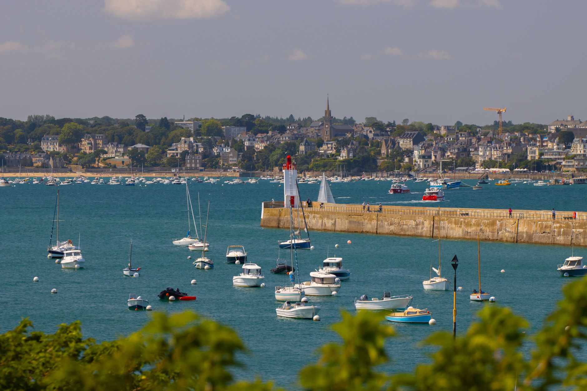 Vue d’un port côtier abrité avec des voiliers et bateaux à moteur amarrés près d’une jetée et d’un phare.
